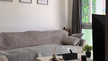 A modern living room with a grey sofa, a white coffee table, and a television. There are framed certificates on the wall and a window with curtains.