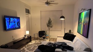 Een woonkamer met een flatscreen televisie, een bank, een salontafel, een lamp en een schilderij aan de muur.