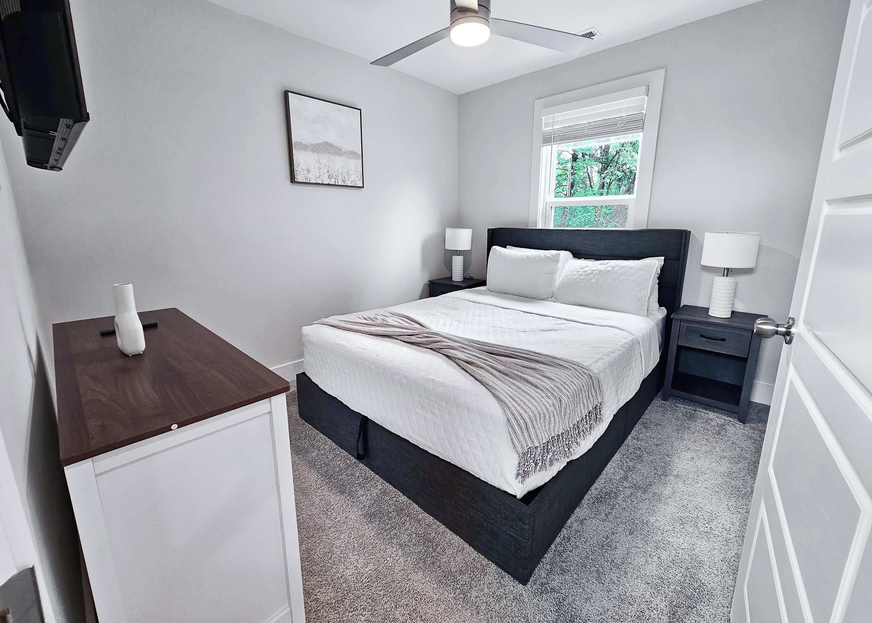 A bedroom with a bed, bedside tables, a nightstand, a window with blinds, a ceiling fan, and a framed picture on the wall.