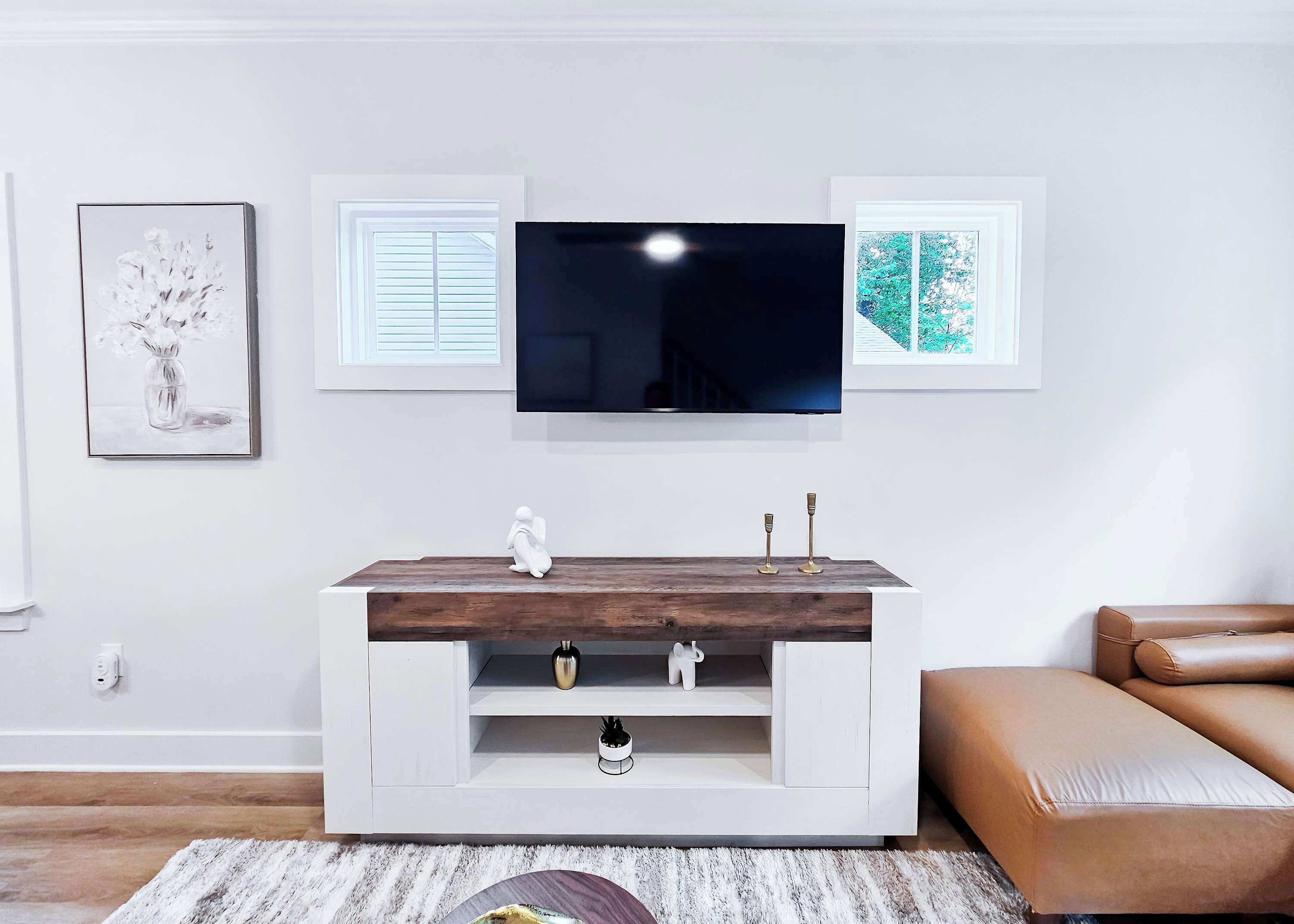 Un moderno salón con un televisor de pantalla plana montado en la pared, una consola blanca con objetos decorativos, un sofá de cuero marrón y cuadros en las paredes.