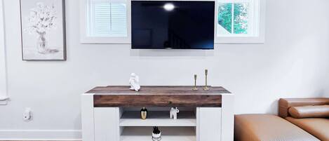 Un moderno salón con un televisor de pantalla plana montado en la pared, una consola blanca con objetos decorativos, un sofá de cuero marrón y cuadros en las paredes.