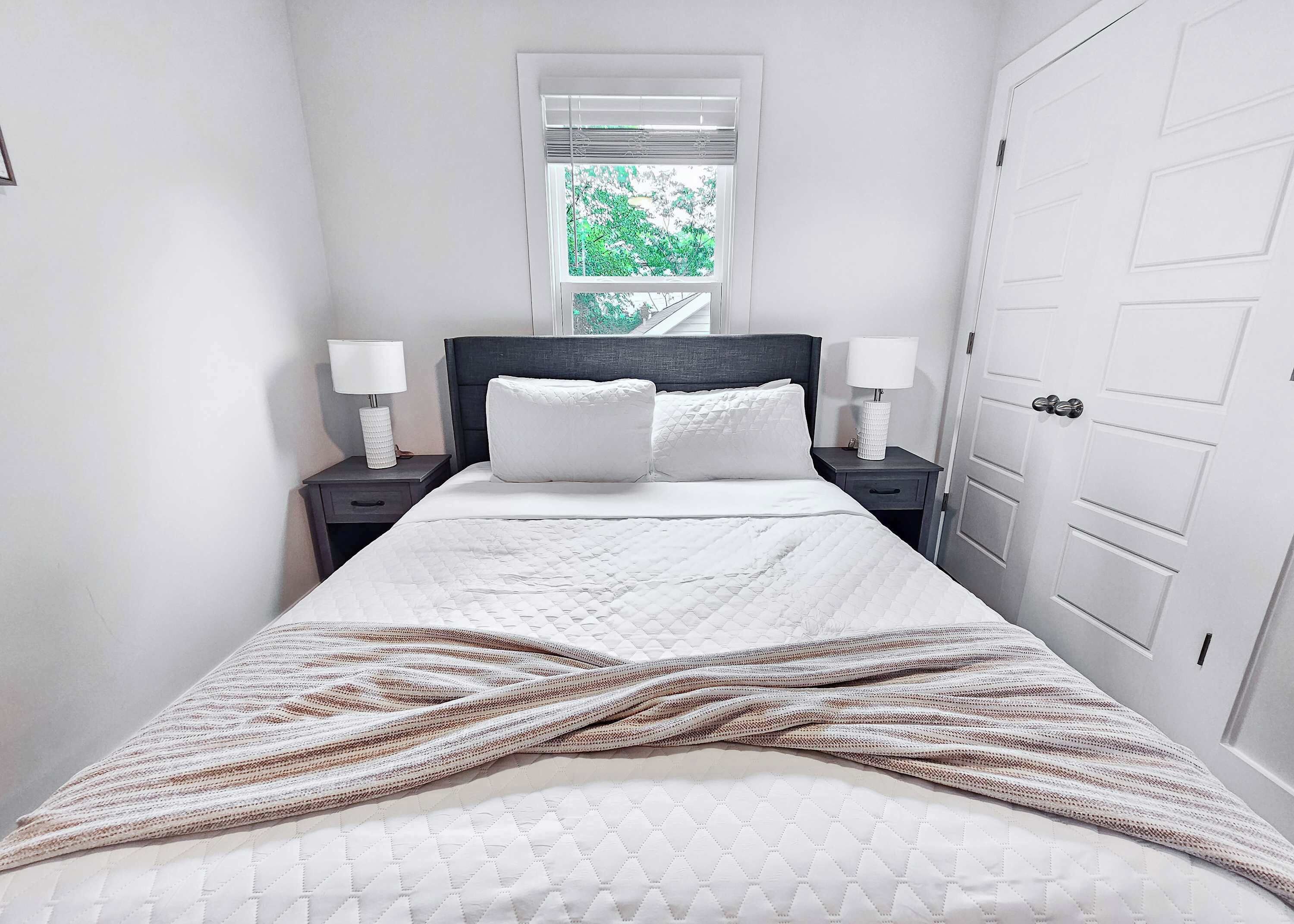 A neatly made bed with a white duvet and pillows, flanked by two bedside tables with lamps, in a room with a window and a white door.