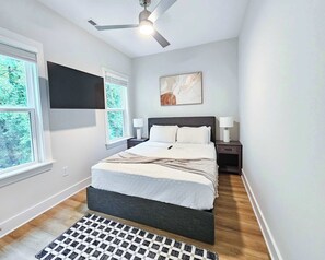 Ein Schlafzimmer mit einem Bett, einem Deckenventilator, einem an der Wand montierten Fernseher und einem Fenster mit Jalousien.