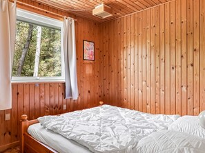 Ein Zimmer mit Holzwänden, einem Bett, einem mit Vorhängen versehenen Fenster und einem an der Wand hängenden Bild.