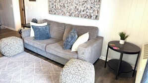En stue med en grå sofa, mønstrede pufs, et lille bord med en plante og et billede på væggen.