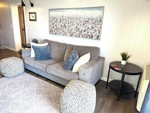 A living room with a grey sofa, patterned ottomans, a small table with a plant, and a painting on the wall. - The Sandy Biscuit!! Oceanfront Corner Condo/Private Beach Access In Windy Hill!! (North Myrtle Beach)
