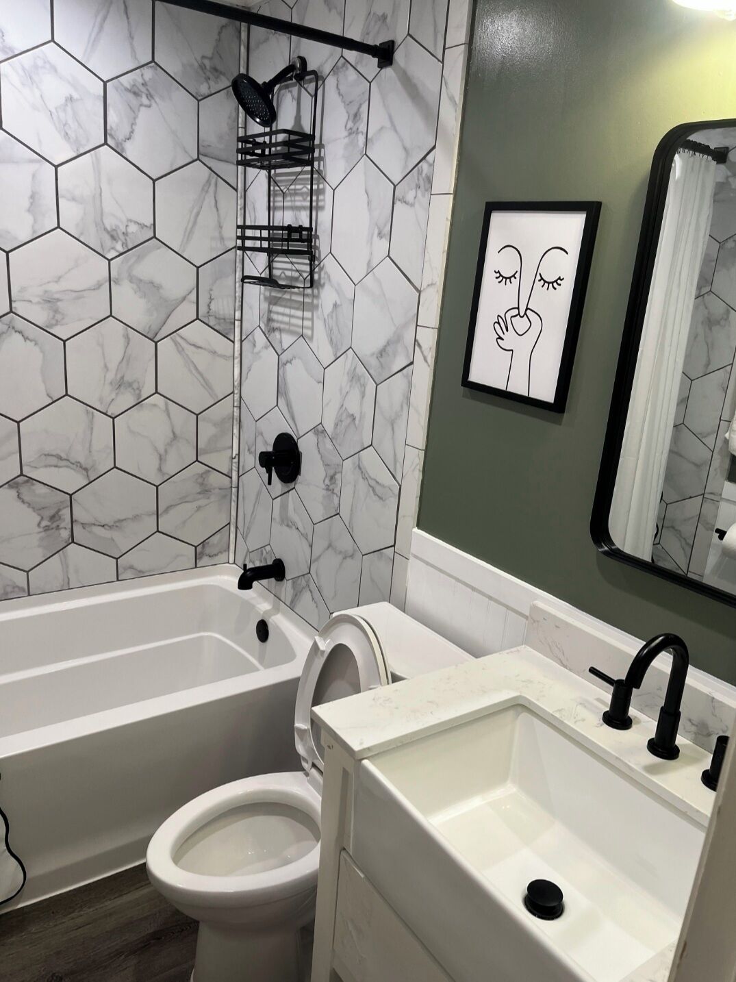 A bathroom with a white bathtub, a toilet, and a sink with a black faucet. There is a shower head on the wall and a framed picture above the sink.