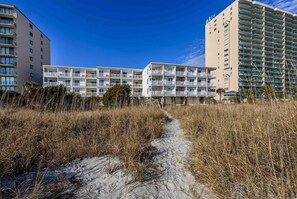 Hotel frente a la playa con varios balcones, un camino de arena que lleva a la entrada y un cielo azul despejado.