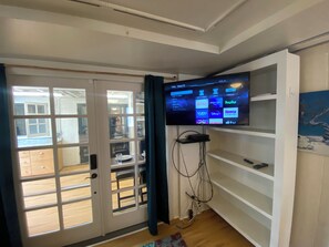 A room with a television mounted on a wall, a door leading to an outdoor area, and a shelf with a remote control.