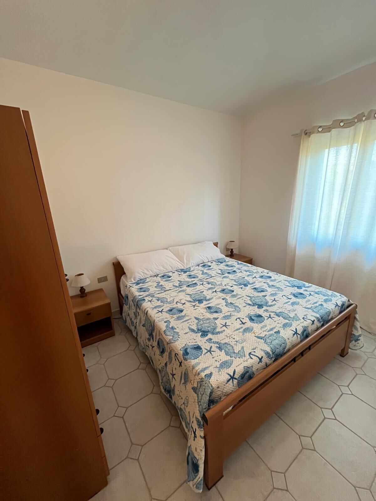 Un dormitorio con cama, mesita de noche y ventana con cortinas.