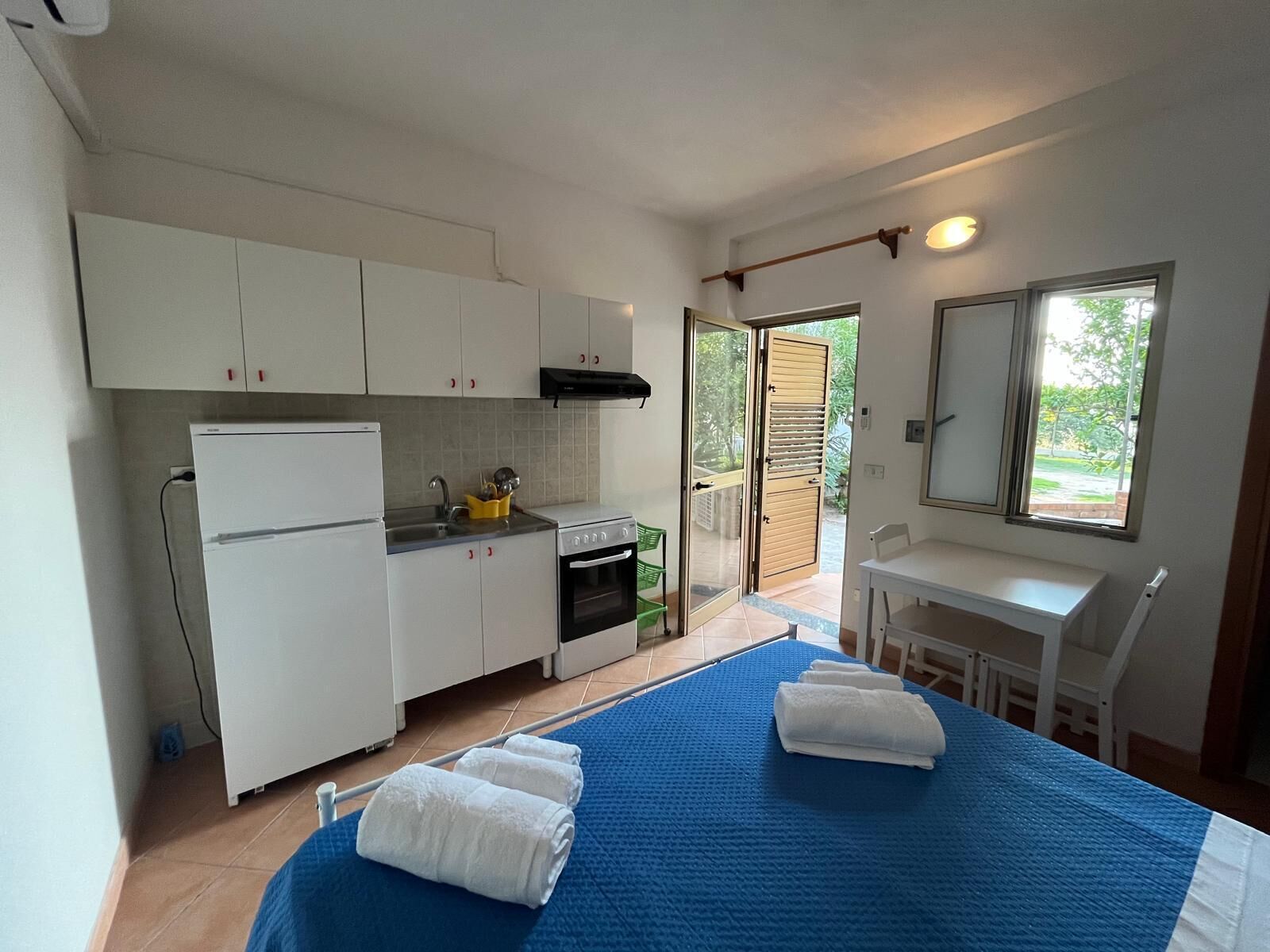 Eine kompakte Küche mit weißen Schränken, einem Herd und einem Spülbecken. Es gibt einen Essbereich mit Tisch und Stühlen, sowie ein Bett mit blauer Bettwäsche.