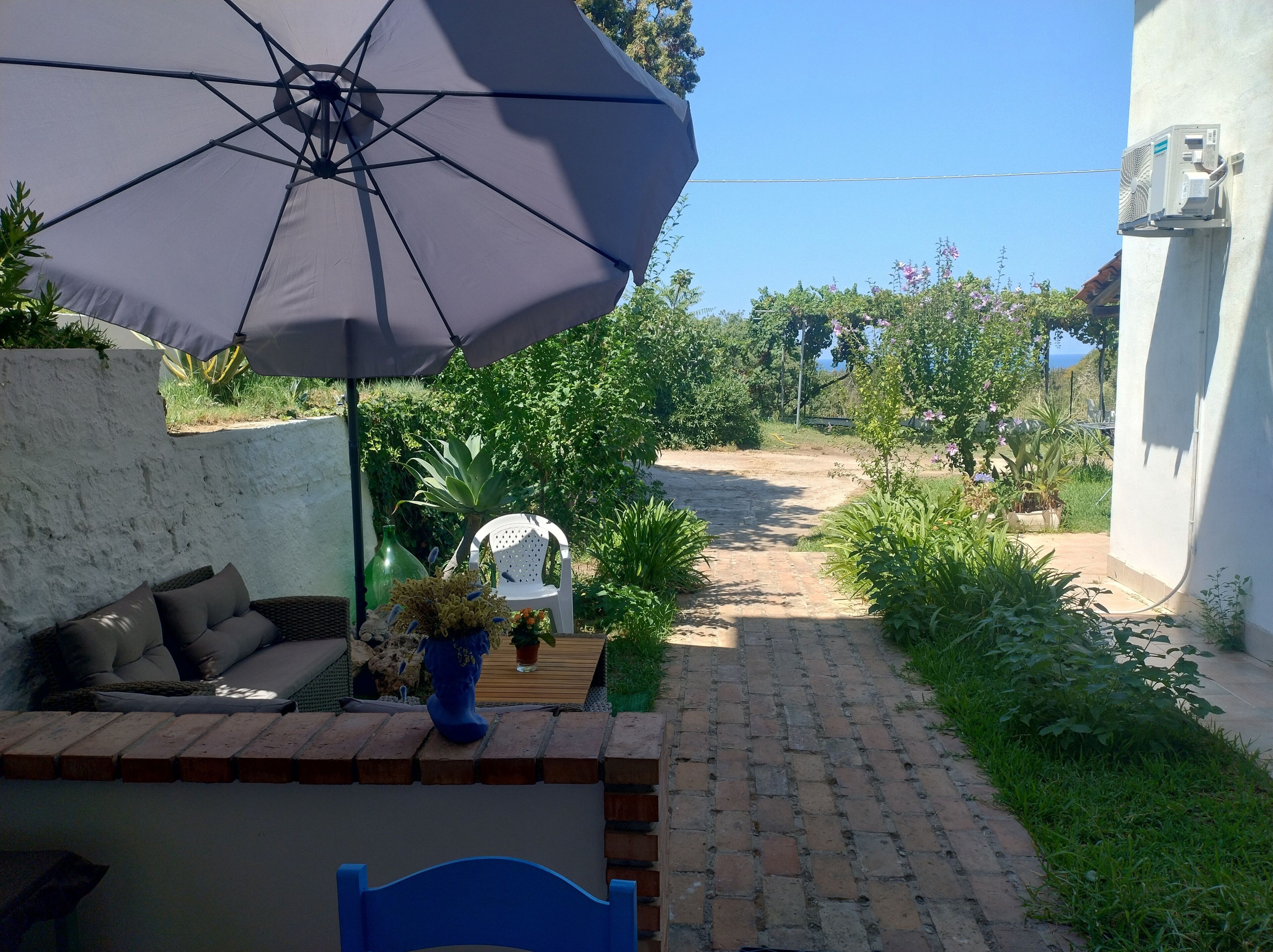 A patio area with a brick pathway, a white chair, a blue chair, a large umbrella, and potted plants.