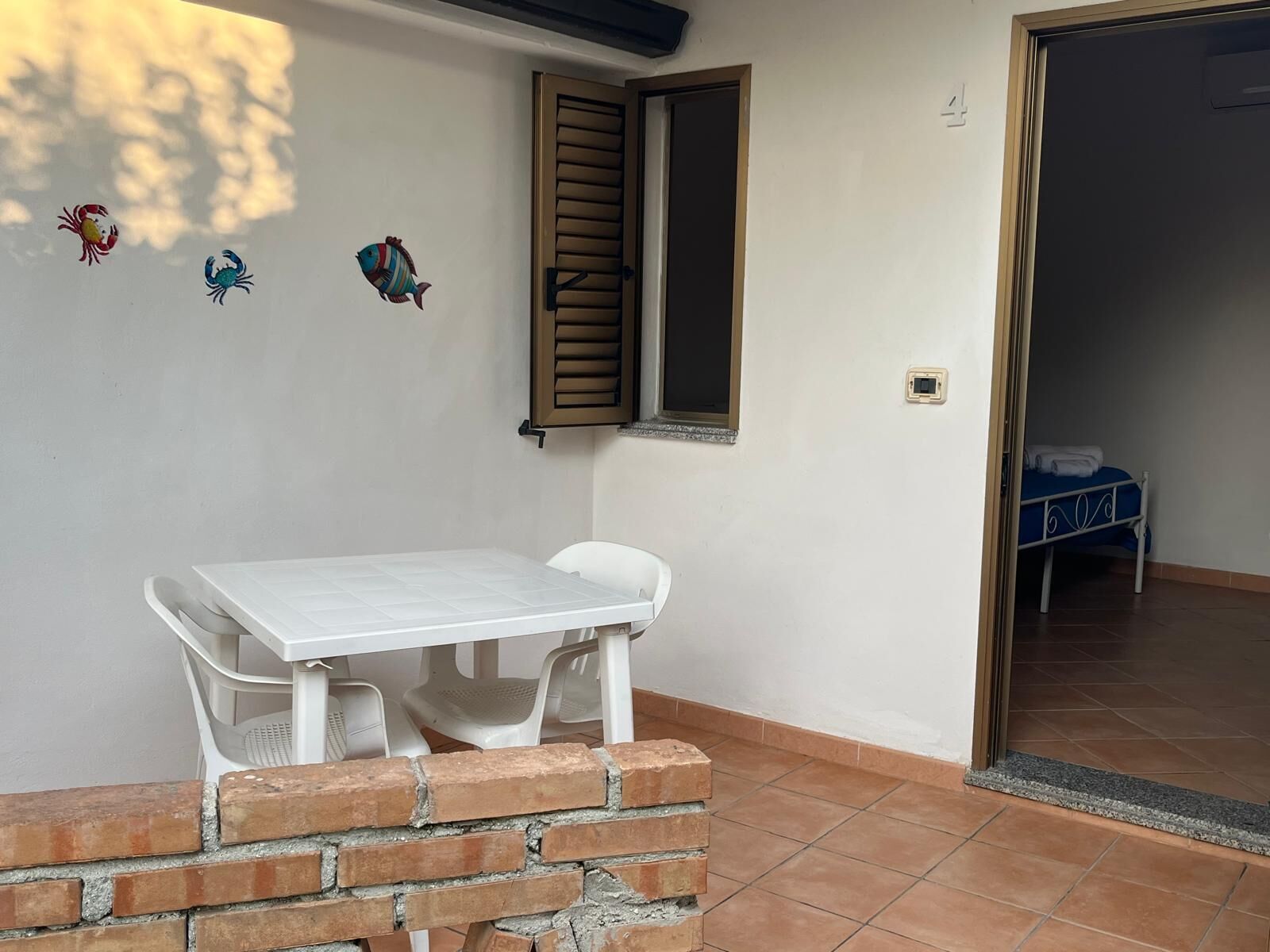 Une pièce avec une table et des chaises blanches, une fenêtre avec des volets et un lit à l’arrière-plan.