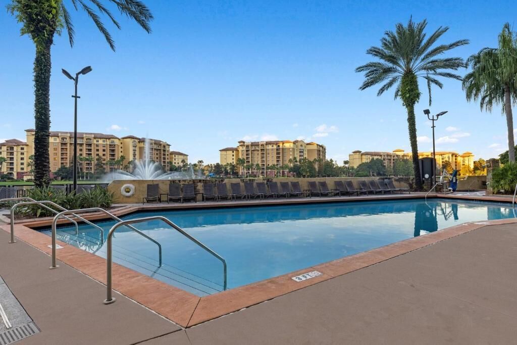 A swimming pool with a water fountain, surrounded by palm trees and lounge chairs, with hotel buildings in the background.
