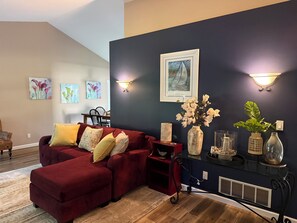 A living room with a red sectional sofa, a dining table with chairs, wall-mounted artwork, and a vase with flowers. - 2BR Charlevoix Getaway - Walk to Lake Michigan Beach - Dog Friendly (Charlevoix)
