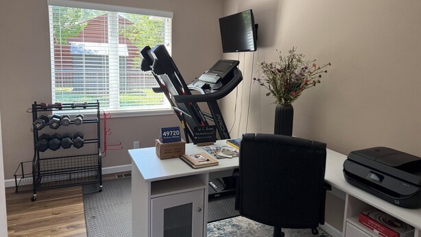 A home office with a treadmill, a desk with a printer, a chair, a small table with a vase, and a window with blinds. - 2BR Charlevoix Getaway - Walk to Lake Michigan Beach - Dog Friendly (Charlevoix)