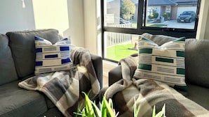 A living room with a sofa, two patterned throw pillows, a blanket, and a view of a residential street through a window. - Bright Kalkallo Home, Easy Melbourne Access (Kalkallo)