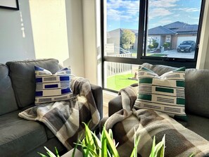 Ein Wohnzimmer mit einer Couch, zwei gemusterten Zierkissen, einer Decke und Blick auf eine Wohnstraße durch ein Fenster.