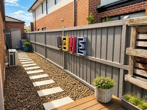 Een buitenruimte met een grindpad, een houten terras, een metalen hek en een potplant.