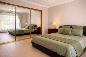 A bedroom with a large mirror, a bed with green bedding, a nightstand with a lamp, and a dresser. - 4BR Home Near Altona Beach · Sleeps 9 · Yard (Altona Meadows)