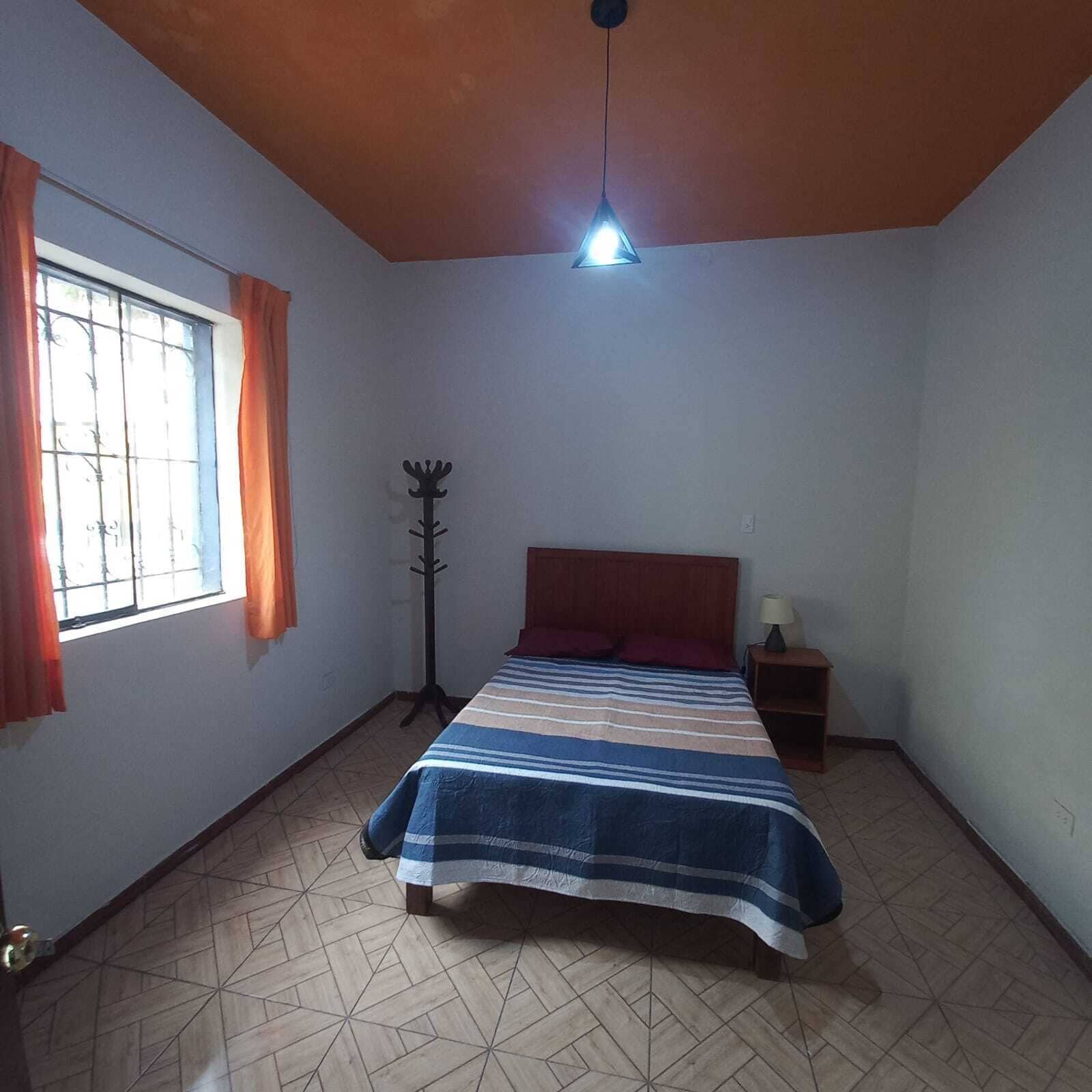 Ein Schlafzimmer mit Bett, Nachttisch, Lampe, Kleiderständer und einem Fenster mit Vorhängen.