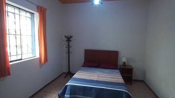 A bedroom with a bed, a nightstand, a lamp, a coat rack, and a window with curtains.
