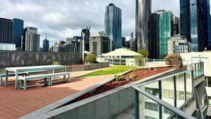 A cityscape with modern high-rise buildings, a rooftop garden with benches, and a glass railing overlooking the city. - Compact Stay with Balcony, Rooftop and Gym (West Melbourne)