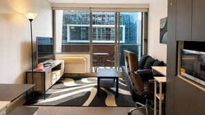 A modern hotel room with a sofa, a desk, a chair, a television, and a large window offering a city view. - Compact Stay with Balcony, Rooftop and Gym (West Melbourne)