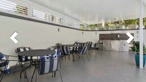 A covered outdoor seating area with tables and chairs, a kitchen area, and a view of greenery. - Location, Location...This seaside unit has it all (Surfers Paradise)