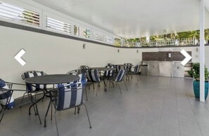 A covered outdoor seating area with tables and chairs, a kitchen area, and a view of greenery. - Location, Location...This seaside unit has it all (Surfers Paradise)