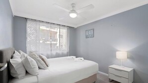 A bedroom with a bed, a nightstand, a lamp, a ceiling fan, and a window with sheer curtains. - Location, Location...This seaside unit has it all (Surfers Paradise)