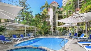 A swimming pool with lounge chairs and umbrellas, surrounded by palm trees and a building with a red roof. - Location, Location...This seaside unit has it all (Surfers Paradise)