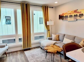 A modern living room with a sofa, a coffee table, and a dining table. There are large windows with curtains, a painting on the wall, and a lamp. - The Nest, TC Downtown + Beach, Rooftop + Garage (Traverse City)