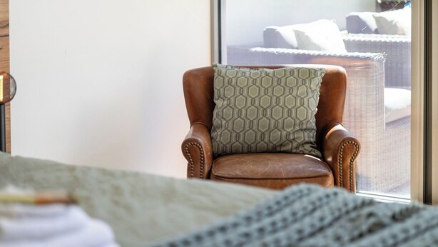 A brown leather armchair with a patterned pillow, positioned in front of a window with a view of a bed and a wicker chair. - Bright and Spacious Penthouse by Parliament House (Forrest)