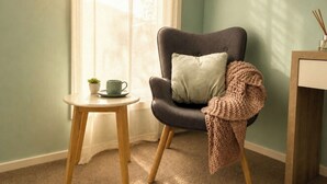 Una habitación con un sillón gris, una mesita redonda con una taza y una planta, y una mesita auxiliar de madera con una vela.