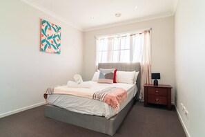 A bedroom with a bed, a bedside table, a lamp, a window with curtains, and a wall art piece. - Spacious Point Cook Retreat w/ Free Parking (Point Cook)