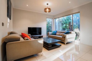A modern living room with a flat-screen TV, two sofas, a coffee table, and large windows with a view of greenery. - Spacious Point Cook Retreat w/ Free Parking (Point Cook)