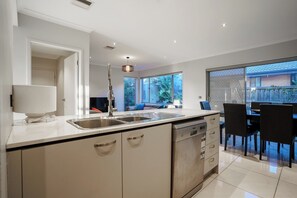 A modern kitchen with a dishwasher, double sink, and a dining area with chairs and a table. - Spacious Point Cook Retreat w/ Free Parking (Point Cook)