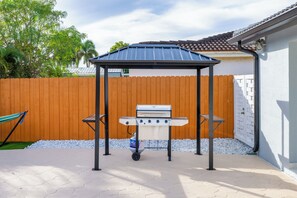 A patio with a barbecue grill under a metal gazebo, surrounded by a wooden fence and a white wall. - Miami Relax Heated Pool Family Getaway (Princeton)