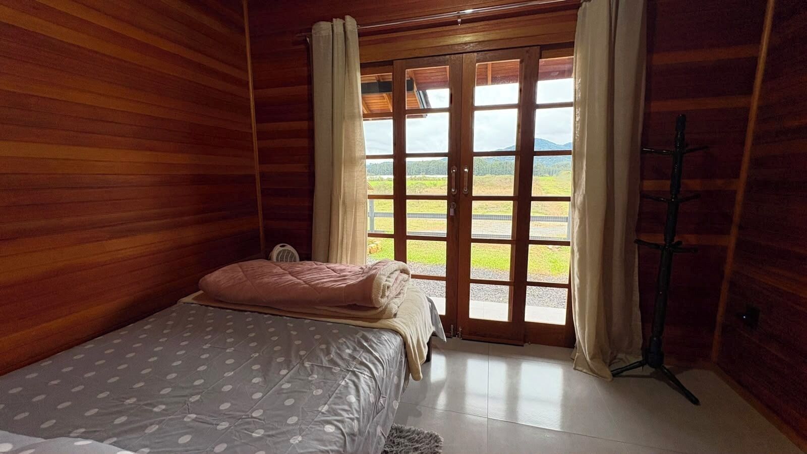 Une pièce en bois avec un lit, un ventilateur sur pied et une vue à travers une porte vitrée.