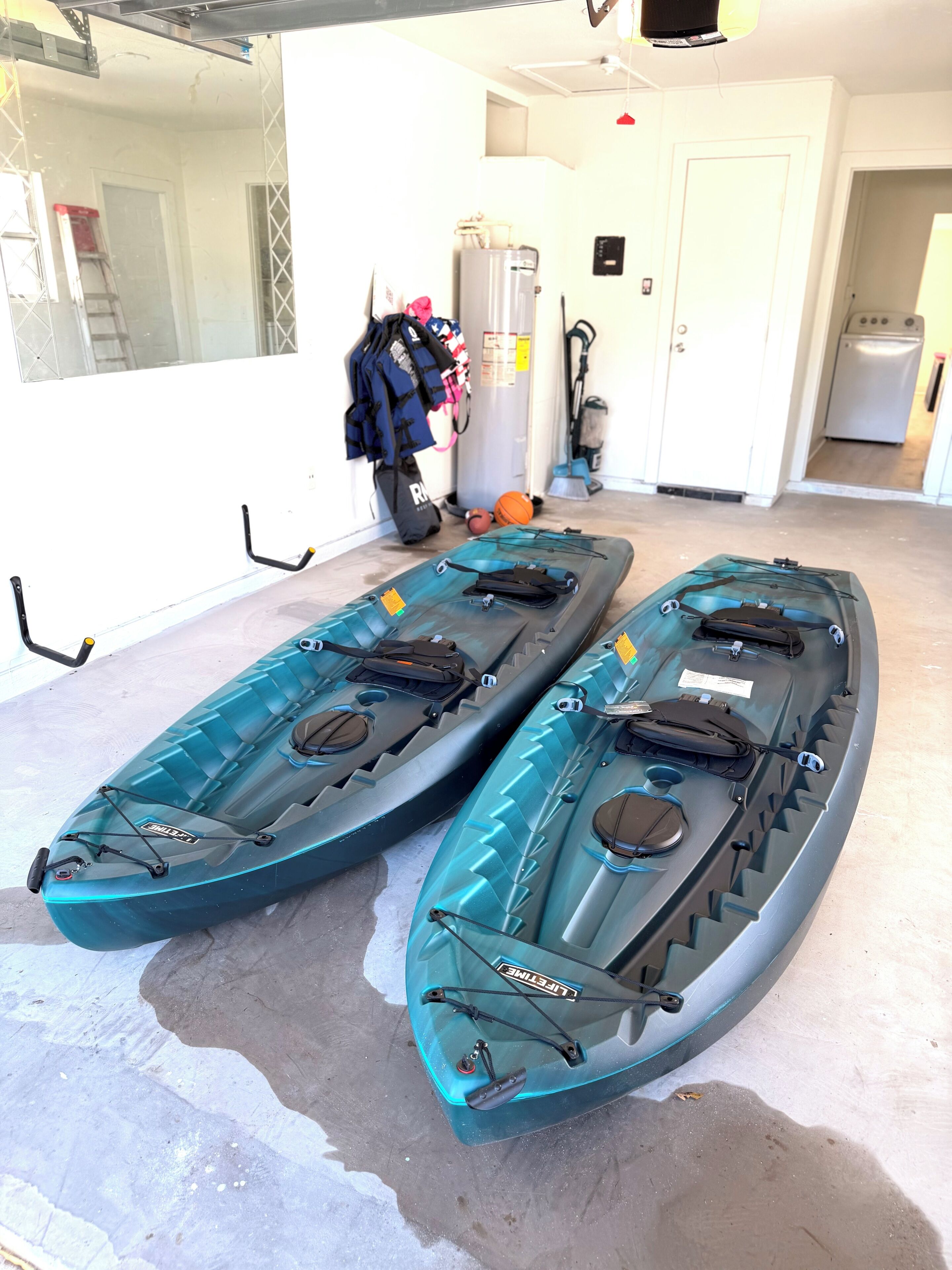 Two kayaks in a garage with a ladder, a basketball, and a vacuum cleaner in the background.