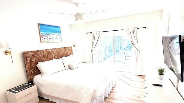 A bedroom with a bed, bedside table, TV, and a window with curtains. -     Coastal Retreat with Pool, Games & Free Kayaks (Sebastian)
