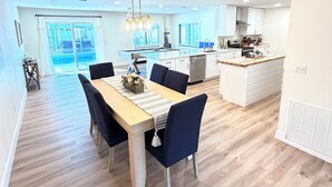 A modern kitchen with white cabinets, a central island, and a dining area with a wooden table and chairs. -     Coastal Retreat with Pool, Games & Free Kayaks (Sebastian)