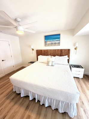 A bedroom with a large bed, a ceiling fan, a painting of a beach, and a nightstand with a lamp. - Coastal Retreat with Pool, Games & Free Kayaks (Sebastian)
