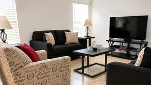 Uma sala de estar com um sofá preto, uma poltrona estampada, uma mesa de centro e uma televisão sobre um suporte.