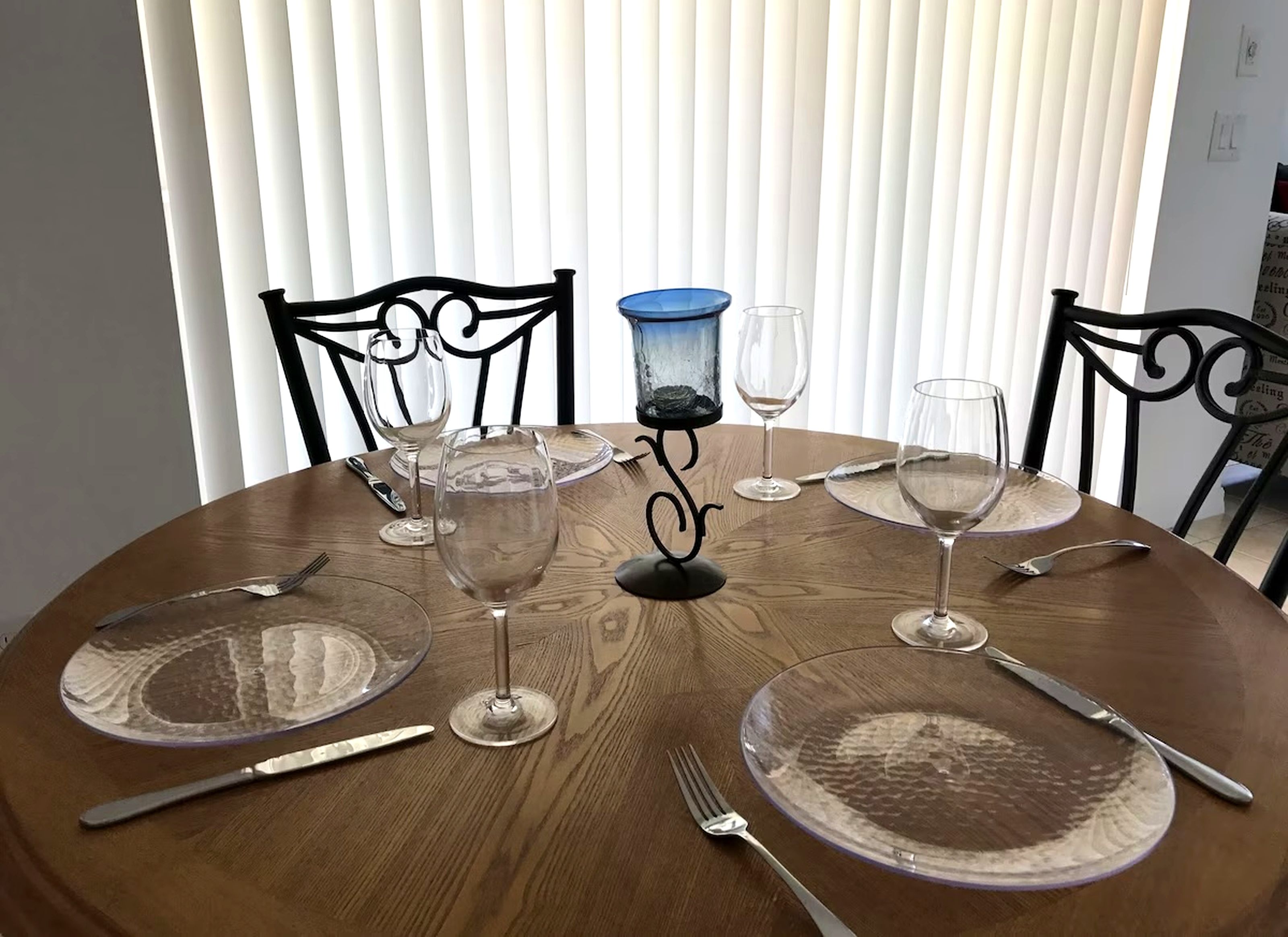Mesa redonda para comer con cristalería transparente, platos y cubiertos, con un fondo de persianas verticales.