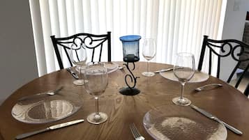 Mesa redonda para comer con cristalería transparente, platos y cubiertos, con un fondo de persianas verticales.