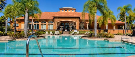 A swimming pool with a tiled deck, surrounded by palm trees and a building with a tiled roof.