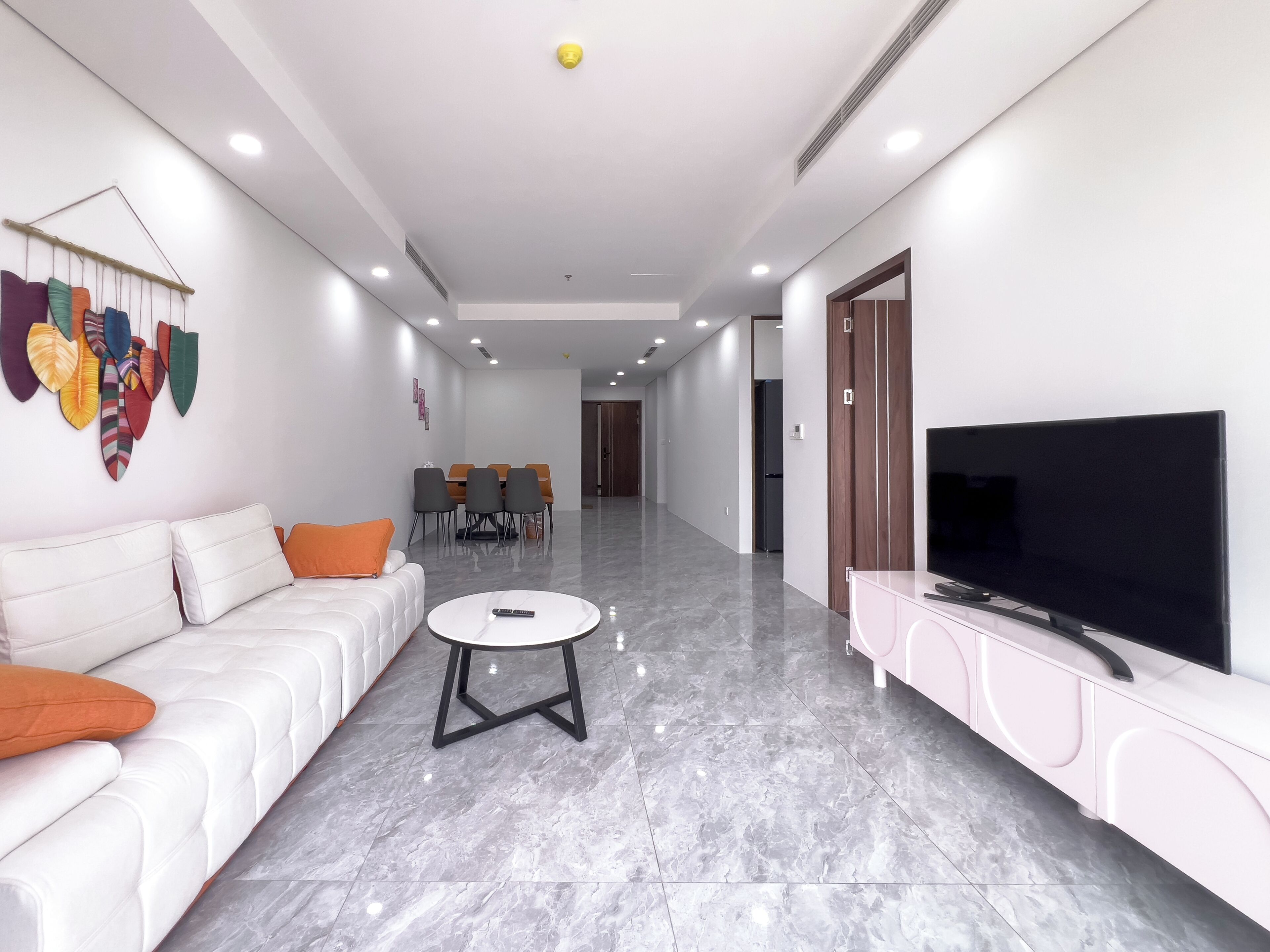 A modern living room with a white sofa, a TV on a pink cabinet, a dining area with chairs, and a hallway leading to a door.
