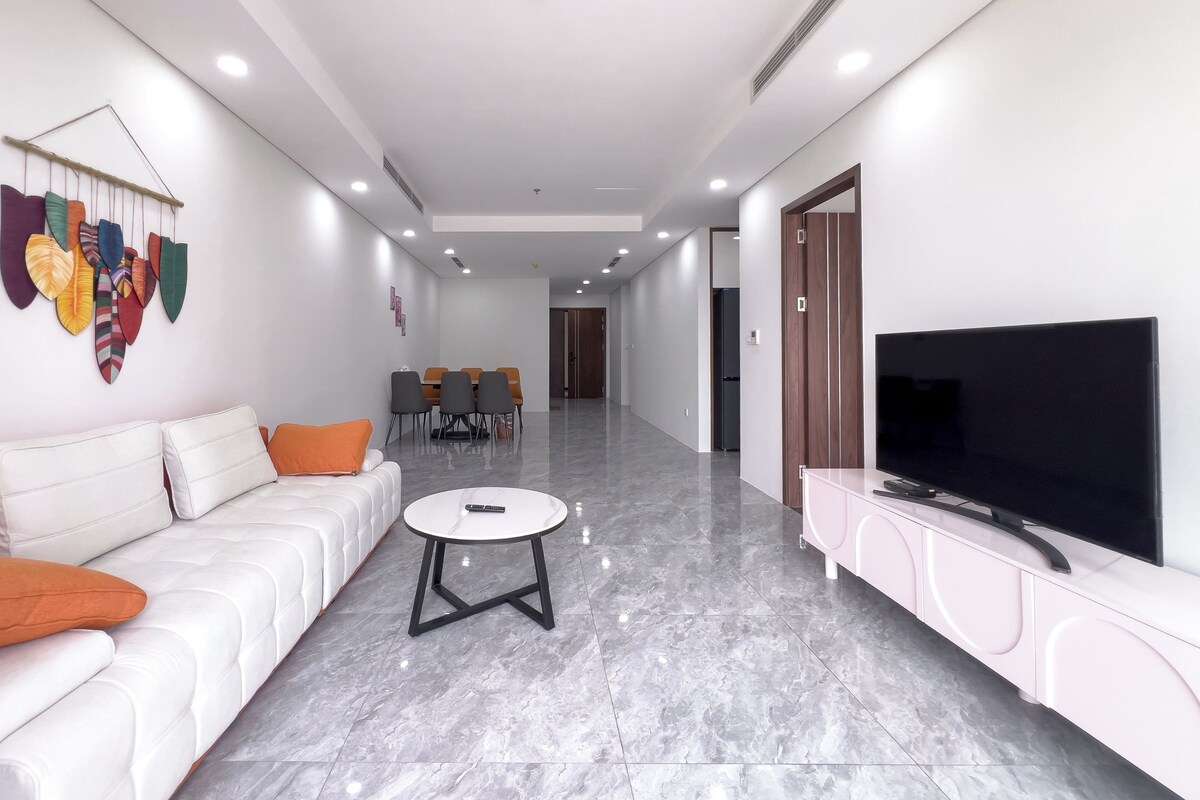 A modern living room with a white sofa, a TV on a pink cabinet, a dining area with chairs, and a hallway leading to a door.